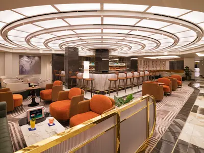 Die Metropolitan Bar an Bord der Norwegian Aqua: Runde Theke mit Barhockern, umgeben von orangen Sesseln und hellen Sofas. Moderne Deckenbeleuchtung und Flaschenregale hinter der Bar dominieren das Bild.