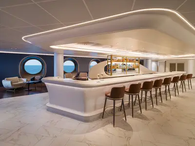Eine elegante Bar-Lounge an Bord der Norwegian Aqua mit runden Fenstern, die den Blick auf das weite Meer freigeben, einer langen Theke und modernen Sitzbereichen.