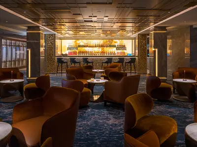 Die elegante Whiskey Bar an Bord der Norwegian Aqua. Gemütliche braune Samtsessel und Tische im Vordergrund. Eine helle, beleuchtete Bar mit Flaschen und Barhockern im Hintergrund. Moderne Deckenstruktur.