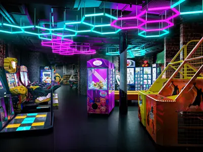 Eine moderne Game Zone an Bord der Norwegian Aqua, beleuchtet von leuchtenden pinken und tuerkisen LED-Lichtern. Zahlreiche Arcade-Automaten, darunter Tanz-, Renn- und Basketballspiele, laden zum Verweilen ein. Der Raum ist ohne Personen.