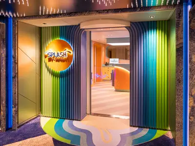 Der farbenfrohe Eingang zur Splash Academy an Bord der Norwegian Aqua mit leuchtenden Lamellen und einem runden, beleuchteten Logo. Im Inneren ist eine Rezeption zu sehen.