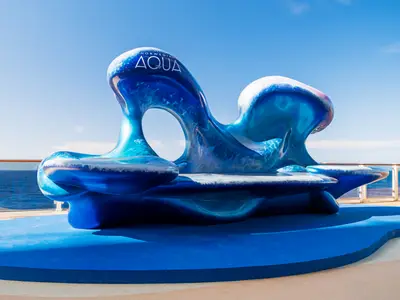 Große, moderne, wellenförmige Skulptur in Blautönen mit 'Norwegian Aqua' Logo auf dem Deck der Norwegian Aqua. Dahinter Meer und Himmel bei Tageslicht. Ein Blickfang an Bord.