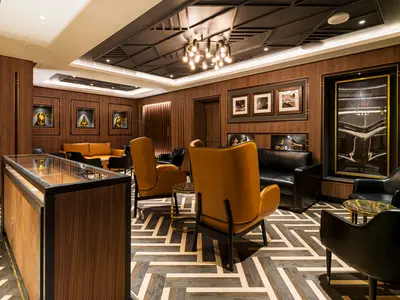 Eine luxuriöse, holzgetäfelte Lounge an Bord der Norwegian Aqua mit bequemen gelben und schwarzen Ledersesseln, einem Kronleuchter und Kunstwerken in Wandnischen.