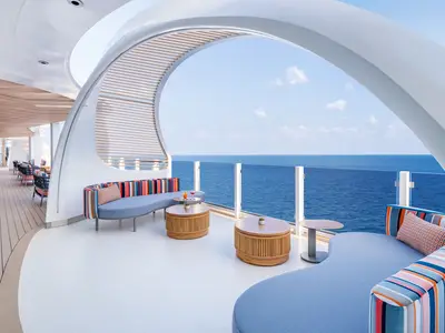 Moderne Aussenlounge der Norwegian Aqua mit bunten Sofas, Holztischen und Cocktails. Weitblick auf das Meer und den Himmel durch eine Glasbrüstung. Organische Designelemente.