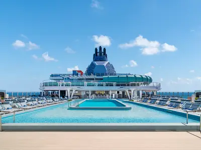 Das Hauptpooldeck der Norwegian Aqua zeigt zwei Pools, leere Liegestühle, eine Poolbar, den Schornstein und eine Wasserrutsche. Blauer Himmel und Meer. Ohne Personen.