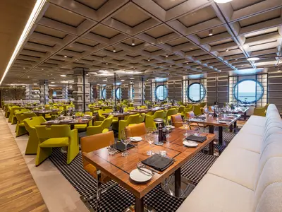 Innenansicht des eleganten Cagney's Steakhouse an Bord der Norwegian Aqua. Der Raum ist mit gelben Stühlen, Holztischen und runden Bullaugen-Fenstern mit Meerblick ausgestattet.