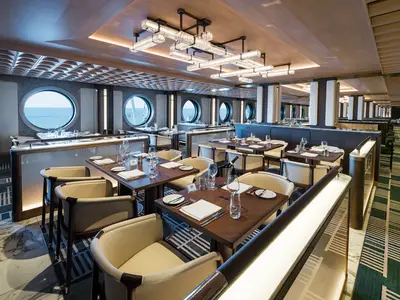 Der Commodore Room der Norwegian Aqua. Ein eleganter Speisesaal mit dunklen Holztischen, beigefarbenen Stühlen, runden Fenstern mit Meerblick und modernen Deckenleuchten.