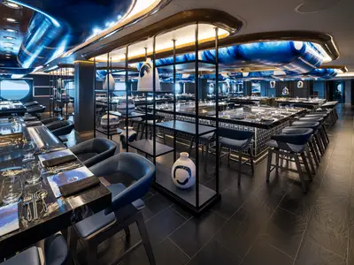 Das moderne Hatsuki Spezialitätenrestaurant an Bord der Norwegian Aqua. Dunkle, gedeckte Tische und Barhocker, blaue Deckenakzente, Vasen und runde Fenster zum Meer.
