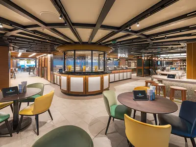 Innenansicht der modernen Indulge Food Hall an Bord der Norwegian Aqua. Mehrere Speisenbereiche wie 'Nudls', 'Just Desserts' und der 'Latin Quarter' sind sichtbar. Bunte Stühle und Tische bieten Platz.