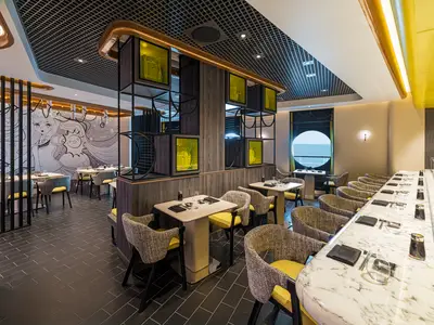 Ein modernes japanisches Spezialitätenrestaurant NamaSushi an Bord der Norwegian Aqua mit dunklem Boden, hellen Tischen, gelb-grauen Stühlen, einer Manga-Wandkunst und runden Fenster mit Meerblick.