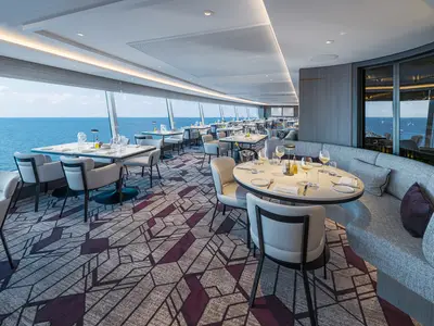 Das Palomar, ein elegantes Spezialitätenrestaurant an Bord der Norwegian Aqua, bietet stilvolle Tische und bequeme Sitzgelegenheiten entlang großer Fenster mit weitem Blick auf das blaue Meer.