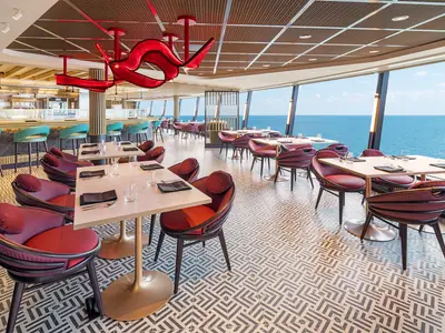 Innenansicht des modernen Spezialitätenrestaurants Sukhothai an Bord der Norwegian Aqua. Mehrere Tische mit roten Stühlen stehen vor grossen Fenstern mit weitem Meerblick. Eine Bar ist im Hintergrund sichtbar, eine rote Skulptur schmückt die Decke.