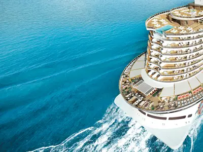Vogelperspektive der Norwegian Aqua auf offener See. Das Bild zeigt das Heck des Schiffs mit mehreren Decks, die mit Lounge-Möbeln, Sonnenschirmen und einem Infinity-Pool ausgestattet sind. Das klare blaue Meer ist mit Wellen zu sehen.