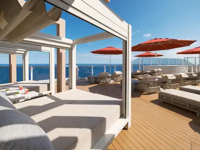 Sonnendeck der Norwegian Aqua: luxuriöse Cabanas, zahlreiche Liegen unter roten Sonnenschirmen. Im Hintergrund Pool und blaues Meer unter klarem Himmel.