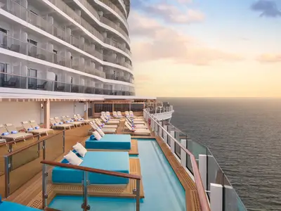 Blick auf das obere Aussendeck der Norwegian Aqua bei Sonnenuntergang. Reihen von Liegestuehlen und blaue Daybeds umrahmen einen schmalen Infinity-Pool. Im Hintergrund sind die Balkonkabinen des Schiffes und das offene Meer sichtbar.