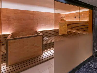 Eine moderne Clay Sauna an Bord der Norwegian Aqua mit orangefarbenen Ziegelwänden und Holzliegen. Warme Lichtbänder beleuchten den Raum. Eine Glaselement trennt den Bereich, rechts ein Infoschild. Zwei gerollte Handtücher liegen auf einer Holzbank.