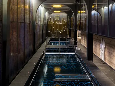 Blick in den eleganten Entspannungsbereich des Mandara Spa an Bord der Norwegian Aqua. Mehrere Pools sind von stilvollen Liegen und einer besonderen Lichtinstallation umgeben. Die Wände zeigen moderne Texturen.