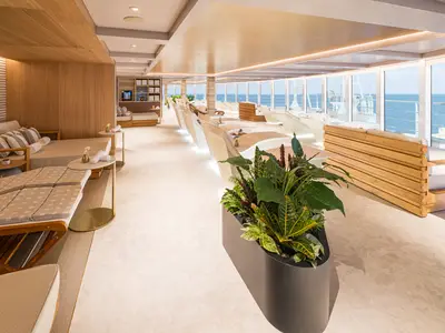 Thermallounge an Bord der Norwegian Aqua. Helle Liegen und Tagesbetten mit Handtüchern. Fensterfront mit Meerblick. Moderner Holz- und Pflanzen-Look.