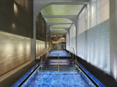 Der elegante Spa-Thermalbereich der Norwegian Aqua. Gestufte blaue Becken, goldene Handläufe, geschwungene Bögen und eine beleuchtete Wasserfallwand.