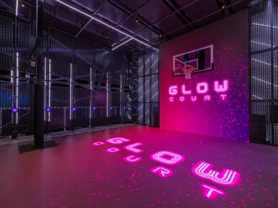 Ein futuristischer Glow Court an Bord der Norwegian Aqua bei Nacht. Das Basketballfeld leuchtet neonpink mit Schriftzügen, umgeben von schwarzen Lochblechwänden und vertikalen Lichtstreifen. Ein Basketballkorb ist an der pink beleuchteten Wand befestigt.
