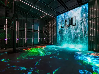 Der futuristische GlowCourt an Bord der Norwegian Aqua. Basketballkorb vor einem digitalen Wasserfall, der Boden leuchtet mit bunten Wellenmustern. Dunkle Gitterwände mit LED-Akzenten. Ohne Personen.