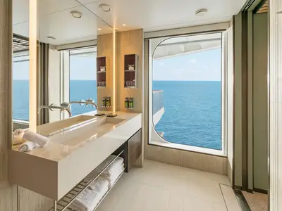 Ein modernes Badezimmer an Bord der Norwegian Aqua mit doppeltem Waschtisch, großem Spiegel und einer breiten Fensterfront, die den Blick auf das offene Meer freigibt. Handtücher liegen bereit.
