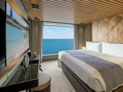 Geräumige Suite an Bord der Norwegian Aqua mit einem großen Bett, einem raumhohen Fenster mit Meerblick und einem großen Flachbildfernseher auf der linken Seite. Die Kabine ist hell und modern eingerichtet.