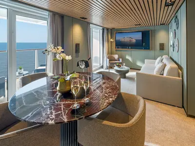 Ein moderner und luxuriöser Wohnbereich in einer Suite an Bord der Norwegian Aqua. Ein Marmortisch mit Sekt und Blumen steht im Vordergrund, dahinter ein Balkon mit Meerblick. Ein Sofa, Sessel und ein Fernseher sind ebenfalls zu sehen.