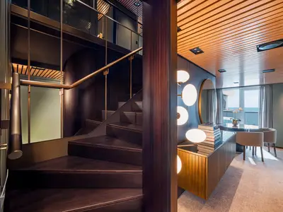 Eine moderne, zweistöckige Suite an Bord der Norwegian Aqua. Eine dunkle Holztreppe führt nach oben, daneben ein heller Wohnbereich mit Meerblick.