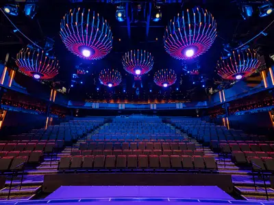The empty AquaTheaterClub on the Norwegian Aqua cruise ship, showcasing multiple tiers of seating under striking, modern, illuminated spherical light fixtures in blue and pink hues.