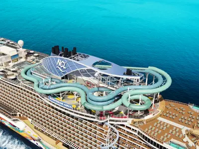 Luftaufnahme der Norwegian Aqua. Riesige Aqua Slidecoaster (blaue Rutschen) dominiert obere Decks. Sonnendecks und blaues Meer sichtbar.
