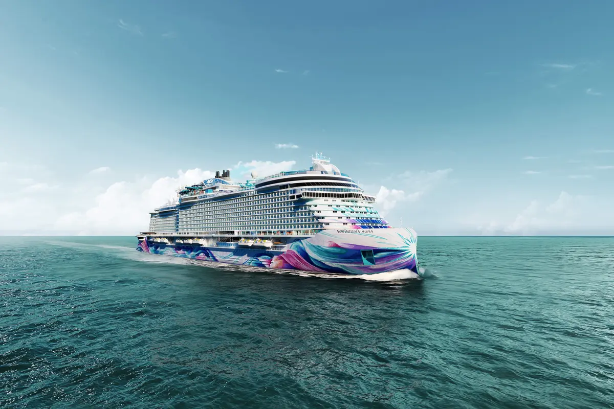 Die Norwegian Aura, ein modernes Kreuzfahrtschiff, fährt auf offener See. Ihr Rumpf zeigt lebhafte Wirbelmuster in Blau, Violett und Pink. Blauer Himmel, leichte Wolken.