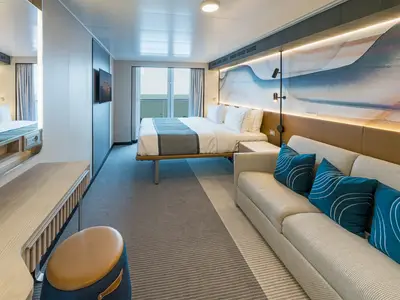 Geräumige Balkonkabine der Norwegian Aura mit einem komfortablen Doppelbett, einem Sofa mit blauen Kissen und direktem Blick auf das Meer vom Balkon.