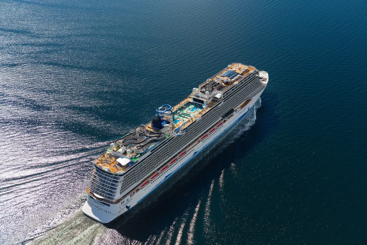 Vogelperspektive des Kreuzfahrtschiffs Norwegian Bliss, das auf blauem Meer fährt. Man sieht die Pools, Wasserrutschen, eine Go-Kart-Bahn und Sonnendecks auf dem Oberdeck.