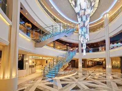 Das moderne Atrium der Norwegian Bliss mit einer blau beleuchteten Glas-Treppe, einem großen Kristall-Kronleuchter, Geschäften und Spielautomaten auf den oberen Decks.