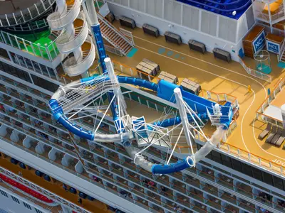 Blick von oben auf die blauen und weissen Wasserrutschen des Aqua Parks an Bord der Norwegian Bliss. Zahlreiche Balkone und ein Joggingpfad sind ebenfalls zu sehen. Mehrere Personen entspannen sich an Deck.