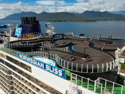 Blick von oben auf die Go-Kart-Rennstrecke an Bord der Norwegian Bliss. Mehrere Karts sind auf der schwarzen Strecke mit blauen und rot-weißen Markierungen zu sehen. Im Hintergrund erstreckt sich ein blaues Meer mit grünen, bewaldeten Bergen unter einem bewölkten Himmel.