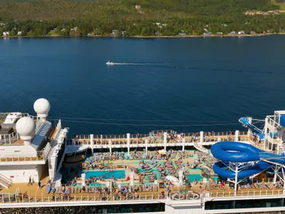Blick von oben auf das Pooldeck der Norwegian Bliss. Gäste entspannen an Pool und Wasserrutschen. Blaues Meer mit Boot und grüne Küste im Hintergrund.