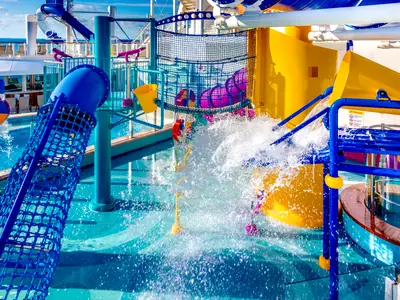Ein lebhafter Kids Aqua Park auf der Norwegian Bliss mit leuchtend blauen und gelben Rutschen, Netzen und spritzendem Wasser. Die Szene zeigt verschiedene interaktive Wasserspiele und bunte Eimer, die Wasser auf die Oberfläche gießen.