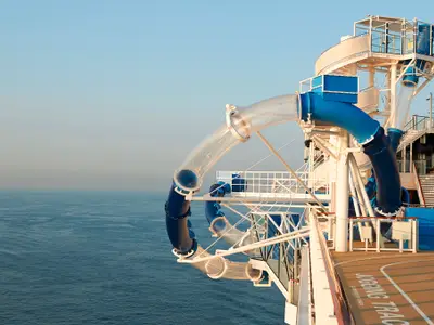 Die aufregende Ocean Loops Wasserrutsche der Norwegian Bliss mit transparenten und blauen Röhren, die sich über dem weiten Meer erstreckt. Ein sonniger Tag an Bord.