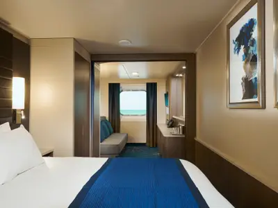Innenansicht einer Aussenkabine an Bord der Norwegian Bliss. Ein Doppelbett im Vordergrund, ein Sofa und ein Fenster mit Meerblick im Hintergrund. Moderne Einrichtung mit blauen Akzenten und Holzelementen.