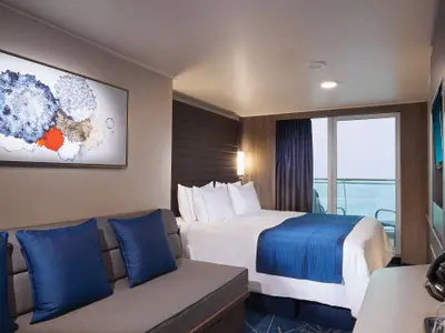 Eine moderne Balkonkabine an Bord der Norwegian Bliss mit großem Bett, Sofa, Balkon mit Meerblick und einem bunten Wandgemälde.