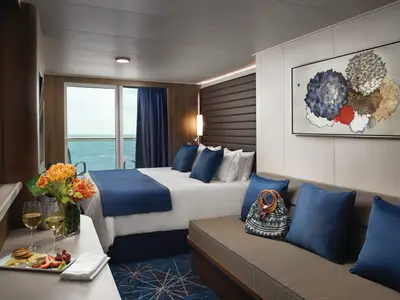 Helle Balkonkabine der Norwegian Bliss mit Doppelbett, blauer Tagesdecke, beigem Sofa und einem Balkon mit Blick auf das Meer. Auf einem Tisch stehen Wein, Käse und Obst.
