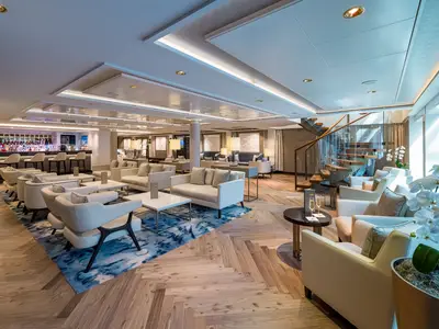 Eine helle, elegante Bar-Lounge an Bord der Norwegian Bliss. Mehrere Sitzgruppen mit beigen Sofas und Sesseln, eine beleuchtete Bar und eine moderne Wendeltreppe sind sichtbar. Der Boden hat ein Fischgrätmuster.