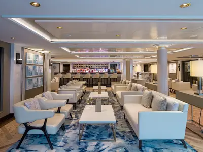 Eine elegante Bar-Lounge an Bord der Norwegian Bliss. Die Szene zeigt helle Sofas und Sessel, kleine Tische und eine beleuchtete Bar mit bunten Flaschen im Hintergrund. Ohne Personen.