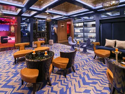 Die elegante Bliss Social Lounge an Bord der Norwegian Bliss mit blauer und goldener Einrichtung, gepolsterten Sitzen und Tischen. Die Raumatmosphäre ist modern und einladend.