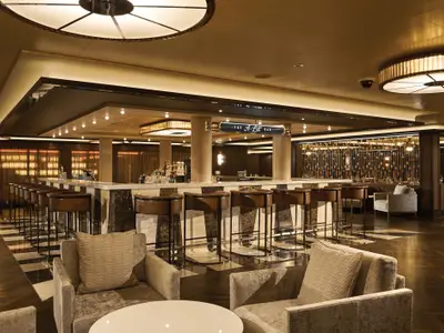 Die schicke A-List Bar auf der Norwegian Bliss mit einer langen Marmortheke und Lederhockern. Warme Beleuchtung, gemütliche Sessel und eine dekorative Wand schaffen eine entspannte Atmosphäre.
