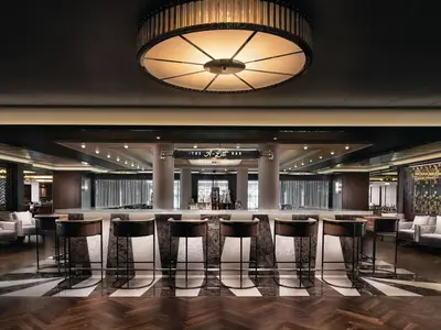 Ansicht der stilvollen A-List Bar an Bord der Norwegian Bliss. Dunkle Holz- und Marmoroberflächen, hohe Barhocker und eine zentrale, runde Deckenleuchte prägen den Raum. Die Bar ist leer, bereit für Gäste.