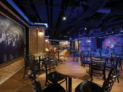 Der Cavern Club an Bord der Norwegian Bliss zeigt eine Bar mit Holztischen, Stühlen und einer beleuchteten Bühne. An den Ziegelwänden hängen große Bilder von Bands und eine bunte Wandbemalung mit Bandnamen.