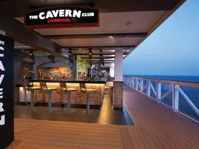 Die Cavern Club Bar an Bord der Norwegian Bliss bei Abendstimmung. Die Bar hat Backsteinoptik und Barhocker. Rechts führt ein offenes Deck mit Geländer zum Meer.